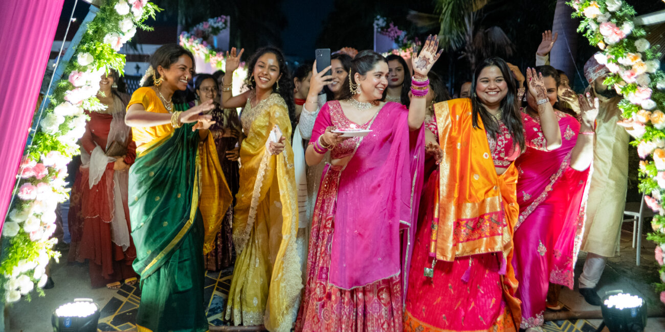 The groom’s sisters dance joyfully, holding silver harathi plates, as they await his arrival during the North Indian wedding procession.