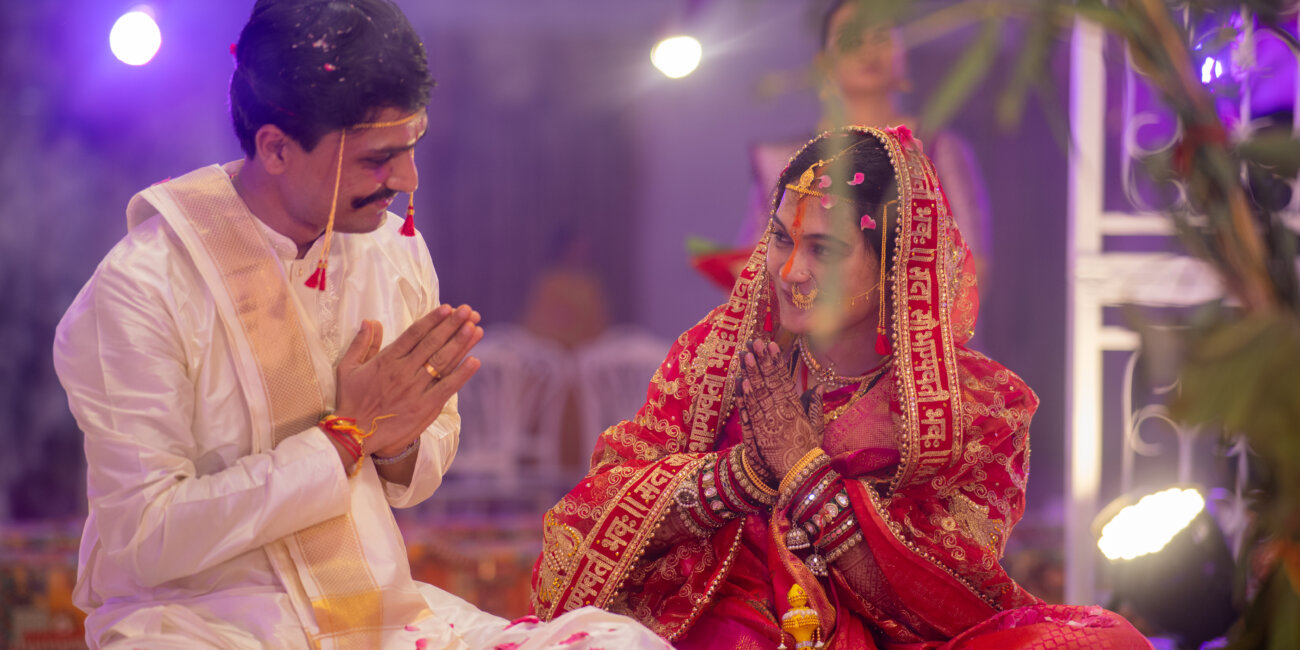 , hands folded in a respectful namaskaram, sharing warm smiles during the wedding ceremony.