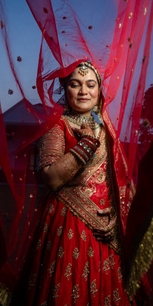 A North Indian bride stands gracefully, eyes closed in a serene expression, her red lehenga’s intricate embroidery glowing in soft golden light.