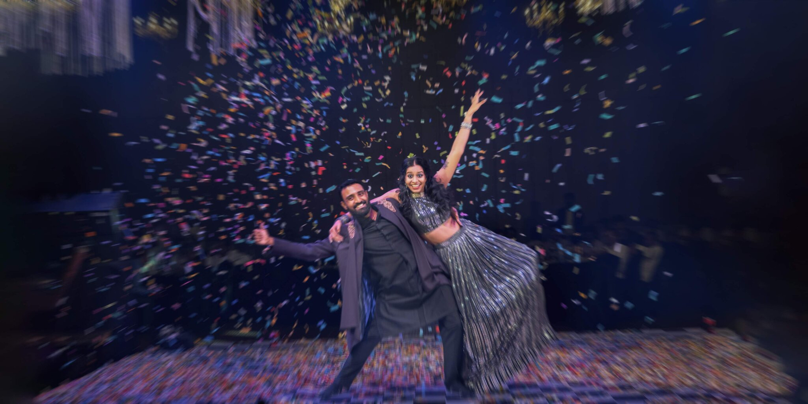 Bride and groom striking a joyful dance pose during the Sangeet, surrounded by colorful lights and festive décor