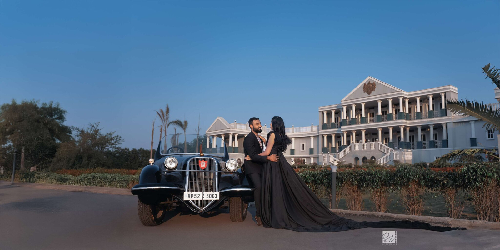 Bride and groom sitting together on a vintage car, with a grand palace standing elegantly in the background, captured in a wide portrait.