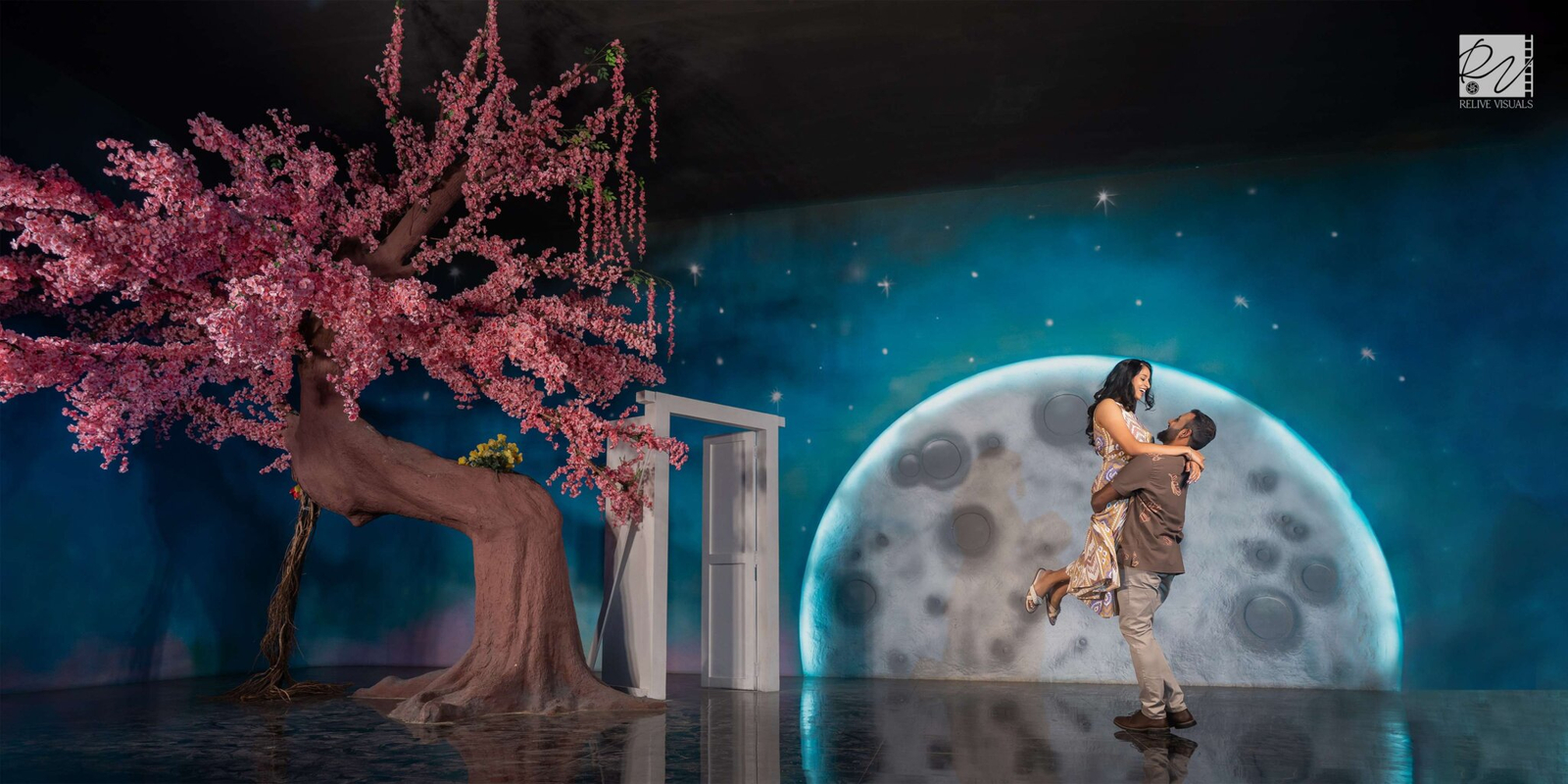 Groom steps back as the bride twirls gracefully under the moonlit sky, framed against a glowing moon backdrop in a wide portrait.